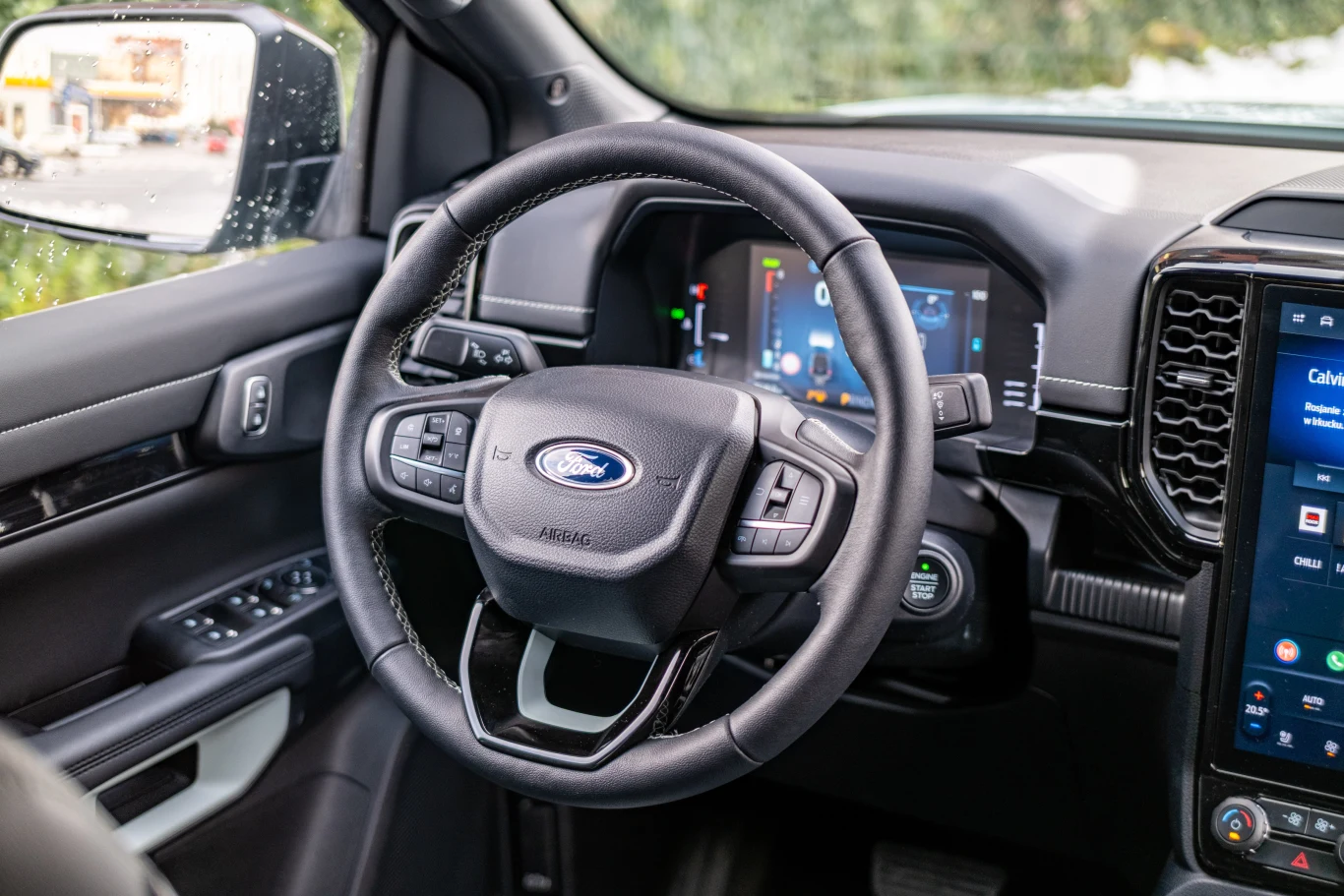 Wnętrze nowoczesnego samochodu Ford z widoczną kierownicą, cyfrowym kokpitem oraz dotykowym ekranem systemu multimedialnego. Detale deski rozdzielczej wykończone wysokiej jakości materiałami, z licznymi przyciskami sterującymi funkcjami pojazdu.