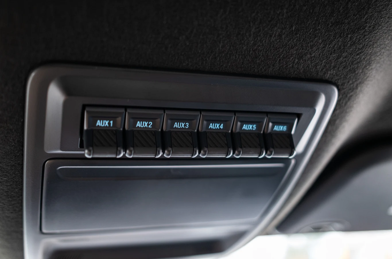 Panel z sześcioma podświetlanymi przełącznikami AUX umieszczony na suficie samochodu, wykonany z tworzywa sztucznego, otoczony czarnym obiciem wnętrza pojazdu.