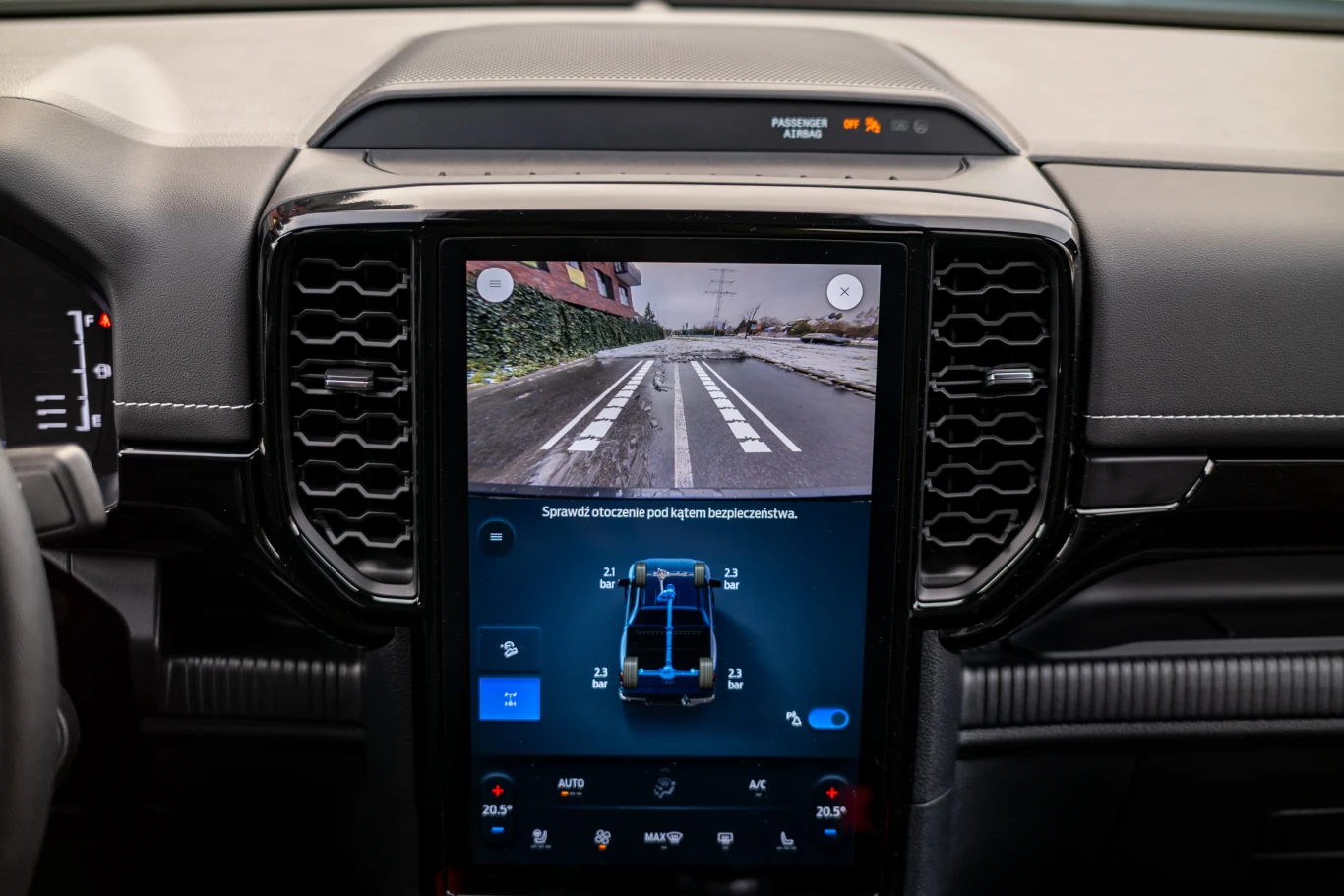 Wnętrze samochodu zdominowane przez dużą centralną konsolę z dotykowym ekranem, na którym wyświetlany jest obraz z kamery cofania oraz informacje dotyczące pojazdu. Widoczne elementy deski rozdzielczej, fragment kierownicy oraz szczegóły systemu multim...