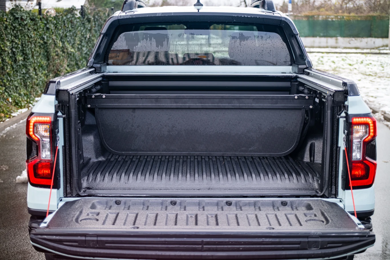 Otwarta przestrzeń ładunkowa pickupa z widoczną klapą, burtami i plandeką, zaparkowanego na asfaltowej drodze w otoczeniu zielonych żywopłotów i śniegu.