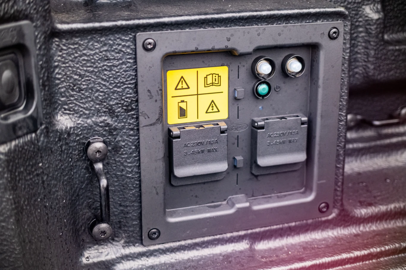 Panel techniczny z dwoma gniazdami elektrycznymi, żółtymi naklejkami ostrzegawczymi oraz metalowymi przyciskami i wskaźnikami świetlnymi, zamontowany na chropowatej metalowej powierzchni.