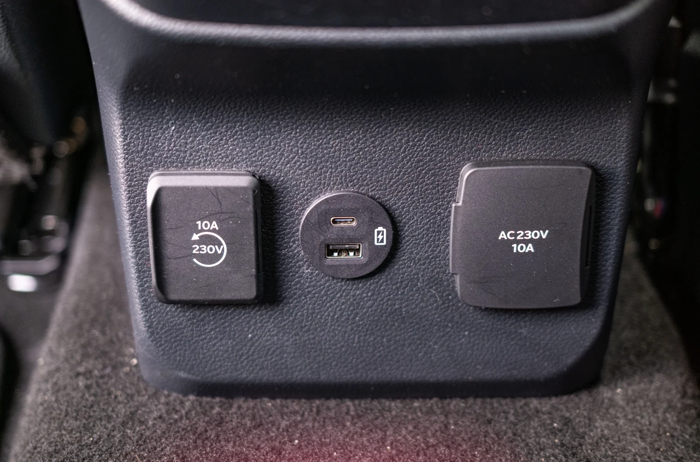 Panel z gniazdem zasilania 230 V oraz portami USB typu A i USB typu C, umieszczony na tle wnętrza samochodu. Obok zamknięte przykrywki oznaczone informacją o natężeniu i napięciu prądu, całość wykonana z tworzywa o fakturze matowej.