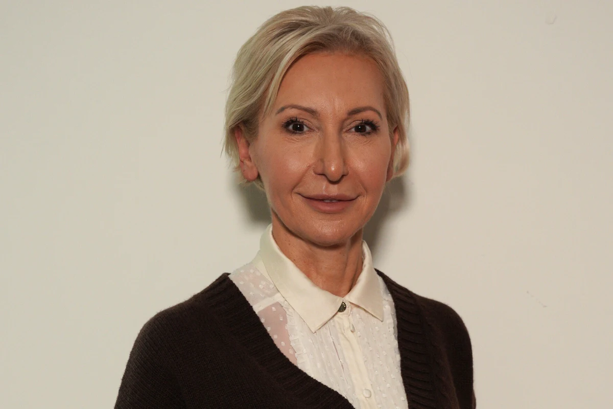 Kobieta o jasnych włosach spiętych z tyłu, ubrana w białą koszulę i ciemny sweter, patrzy prosto w obiektyw na jasnym tle.