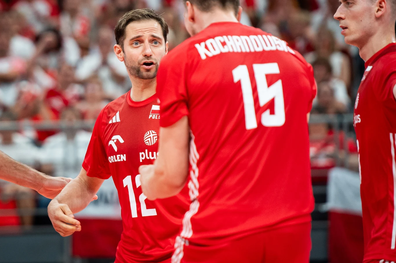 Grzegorz Łomacz w narodowych barwach z numerem 12 Polscy siatkarze w czerwonych strojach w trakcie rozmowy podczas meczu na hali, w tle widoczni kibice w biało-czerwonych barwach.