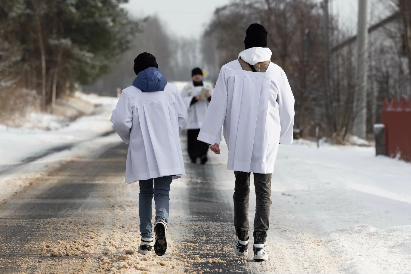 Ministranci towarzyszą księżom podczas kolęd Dwie osoby ubrane w białe fartuchy idą zaśnieżoną drogą, widoczne sylwetki z tyłu, w oddali kolejna osoba także w białym fartuchu, otoczenie zimowe z pokrytą śniegiem nawierzchnią i drzewami po bokach.