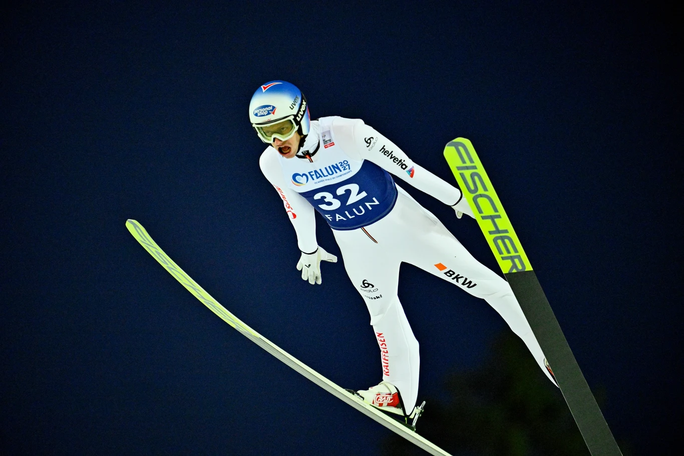 Zawodnik w białym kombinezonie narciarskim podczas skoku na nartach, widoczny numer startowy 32 oraz narty z widocznym logo producenta Fischer, głębokie ciemnoniebieskie tło podkreślające dynamikę momentu.