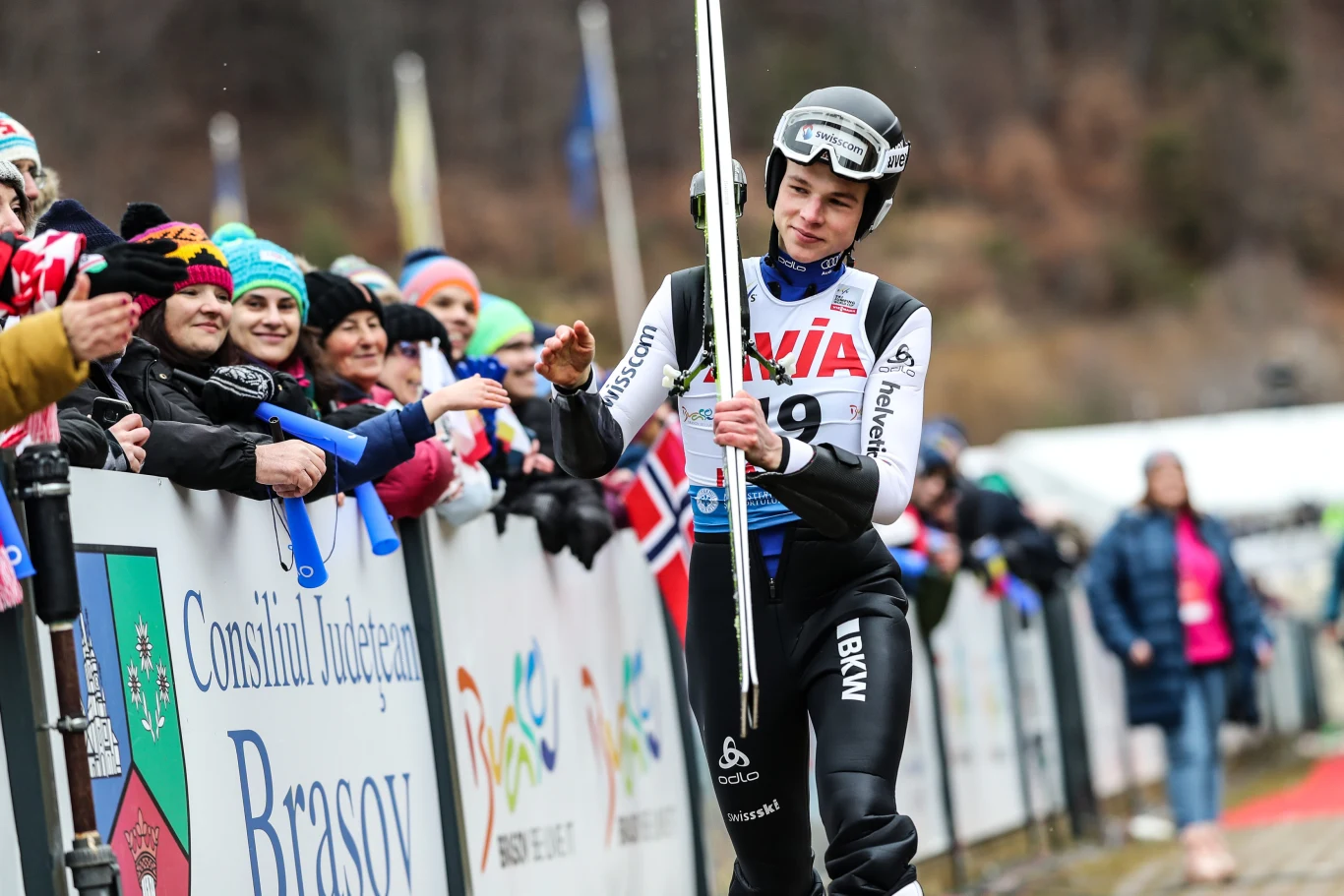 Zawodnik w stroju narciarskim po zakończonym skoku oddaje piątkę kibicom stojącym przy barierce. Wokół sportowca zebrana jest grupka entuzjastycznych fanów w różnobarwnych czapkach, na tle banerów i flag. W tle rozmyty krajobraz ze wzgórzami.