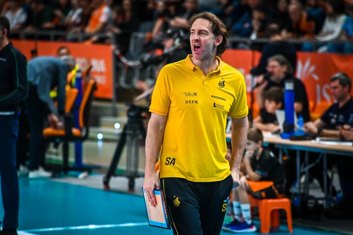 Trener w żółtej koszulce stoi na boisku sportowym, trzymając w ręce notatnik. W tle widownia i osoby siedzące przy stole sędziowskim, a atmosfera wskazuje na trwający mecz siatkówki.