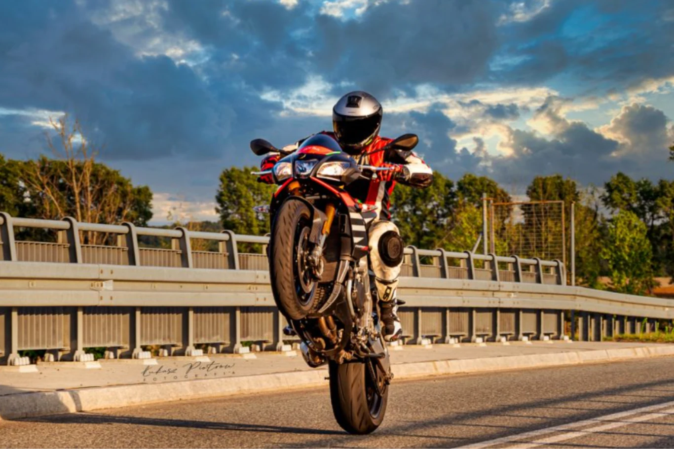 Motocyklista w kolorowym kombinezonie wykonuje efektowny wheelie na sportowym motocyklu na pustej drodze, w tle metalowe barierki i zieleń drzew pod zachmurzonym niebem.