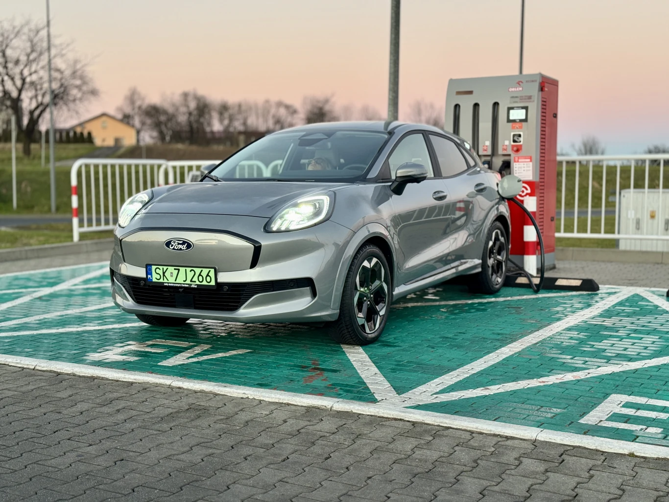 7 na 10 aut służbowych musi być elektrycznych. UE ma nowe limity dla Polski Srebrny samochód marki Ford znajduje się na stacji ładowania pojazdów elektrycznych, na wyznaczonym zielonym miejscu parkingowym, w otoczeniu otwartej przestrzeni i niewielkiej infrastruktury.