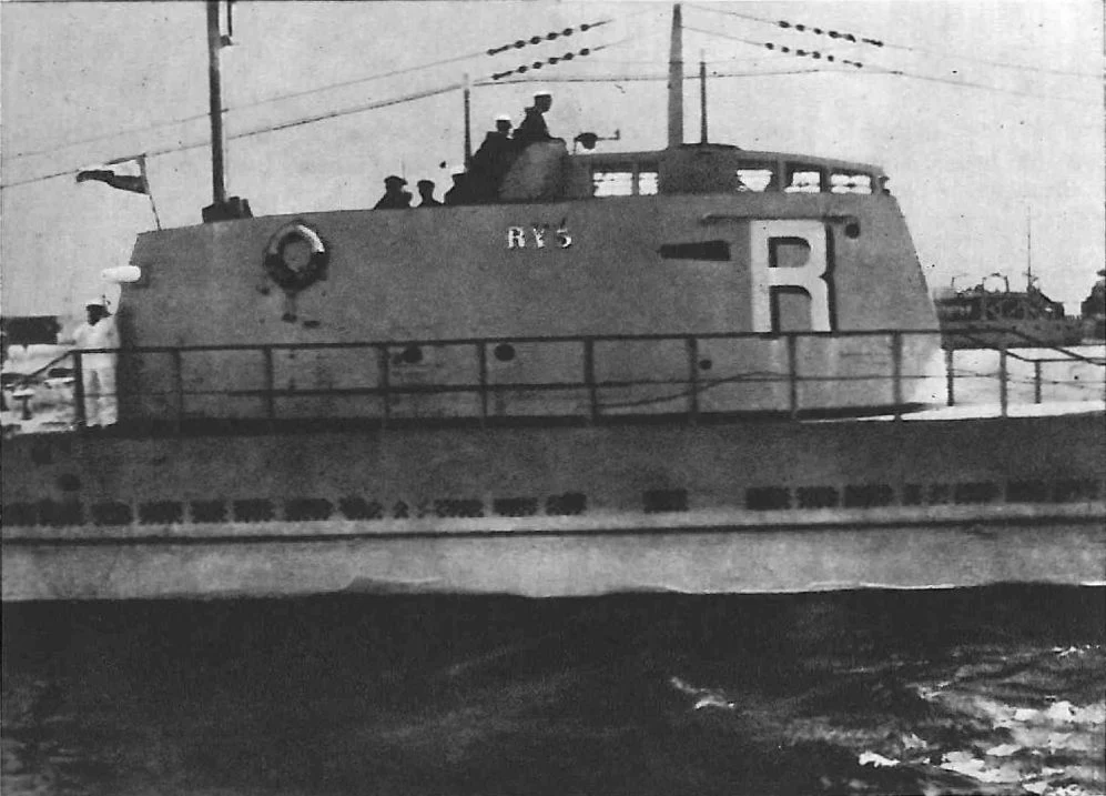 Okręt podwodny z widocznym mostkiem, na którym stoi kilku marynarzy. Na burcie widoczny duży znak 'R' oraz oznaczenie jednostki. Okręt znajduje się na wodzie, w tle zarysy innych statków lub infrastruktury portowej.