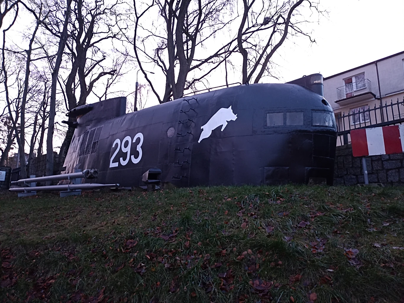 Wypukła, ciemna wieża okrętu podwodnego z numerem 293 i namalowanym symbolem delfina stoi na trawie w otoczeniu drzew, z budynkiem mieszkalnym i metalowym ogrodzeniem w tle.