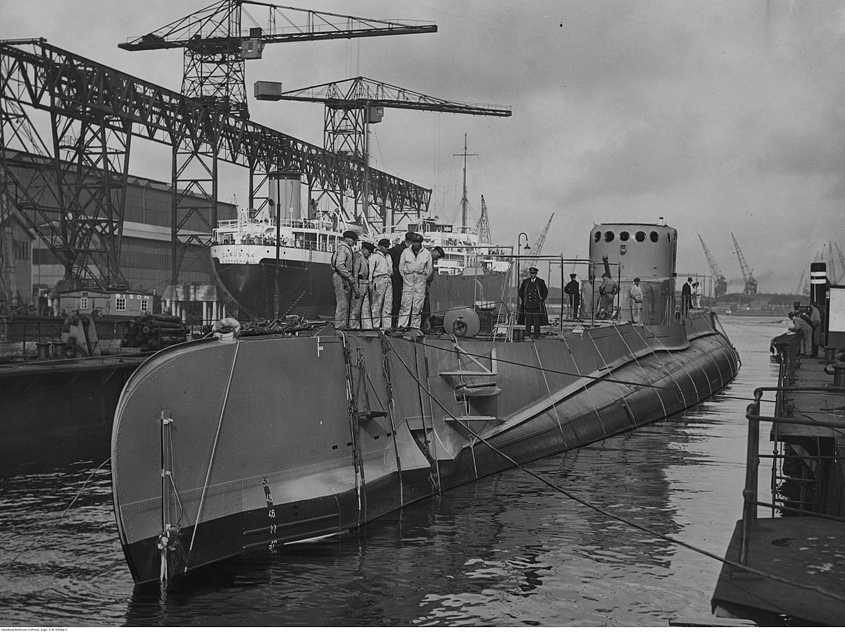 Duży okręt podwodny przycumowany do brzegu w stoczni, na pokładzie stoi kilku mężczyzn w roboczych ubraniach, w tle widać dźwigi i inne jednostki pływające oraz zabudowania przemysłowe