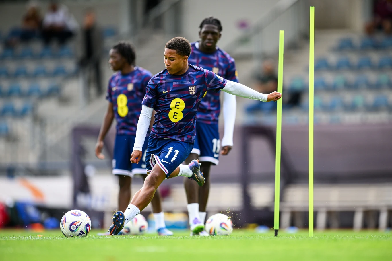 Ajay Tavares Młody piłkarz w stroju treningowym wykonuje drybling piłką pomiędzy żółtymi tyczkami na boisku, w tle znajdują się inni zawodnicy przygotowujący się do ćwiczeń oraz puste trybuny stadionu.