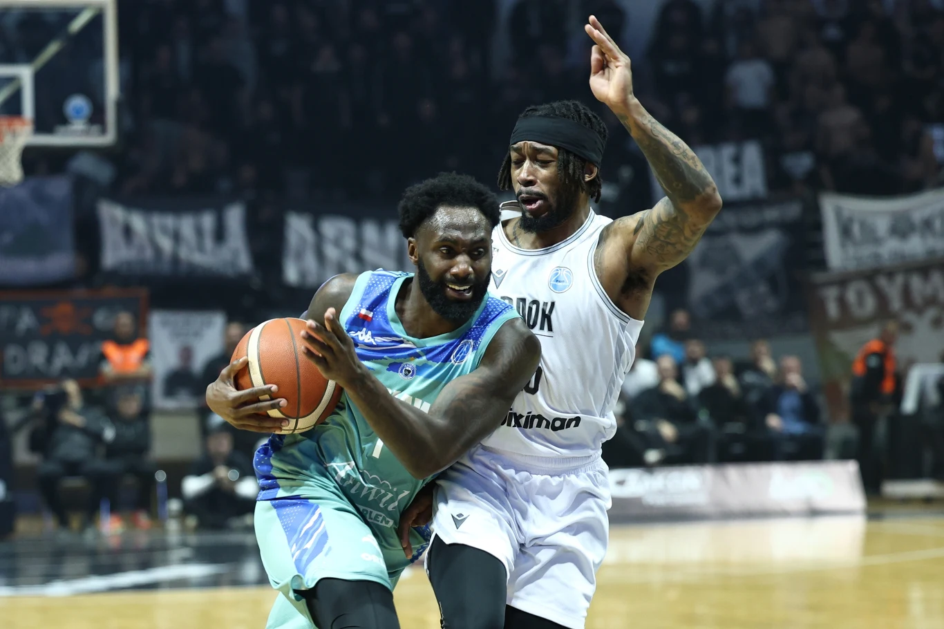 Zawodnik w turkusowym stroju dynamicznie prowadzi piłkę, jednocześnie będąc agresywnie bronionym przez koszykarza w białym stroju, w tle widać trybuny i kibiców.