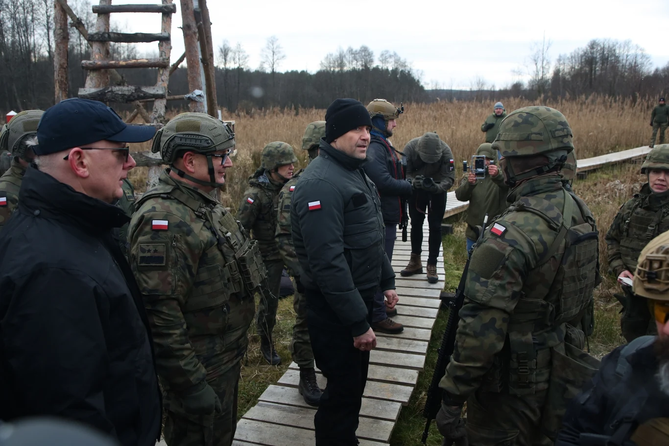 Grupa żołnierzy w mundurach oraz wojskowym wyposażeniu rozmawia z cywilnym mężczyzną na drewnianej kładce w terenie leśno-polnym, w tle widoczna trawiasta roślinność i kilka osób dokumentujących spotkanie.