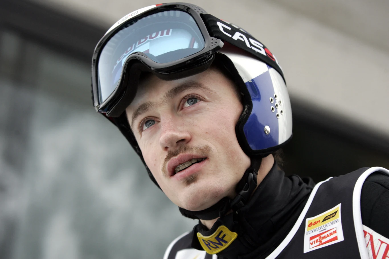 Młody mężczyzna w stroju narciarskim, z kaskiem i goglami, patrzy w górę; na jego kombinezonie widoczne są emblematy sponsorów.