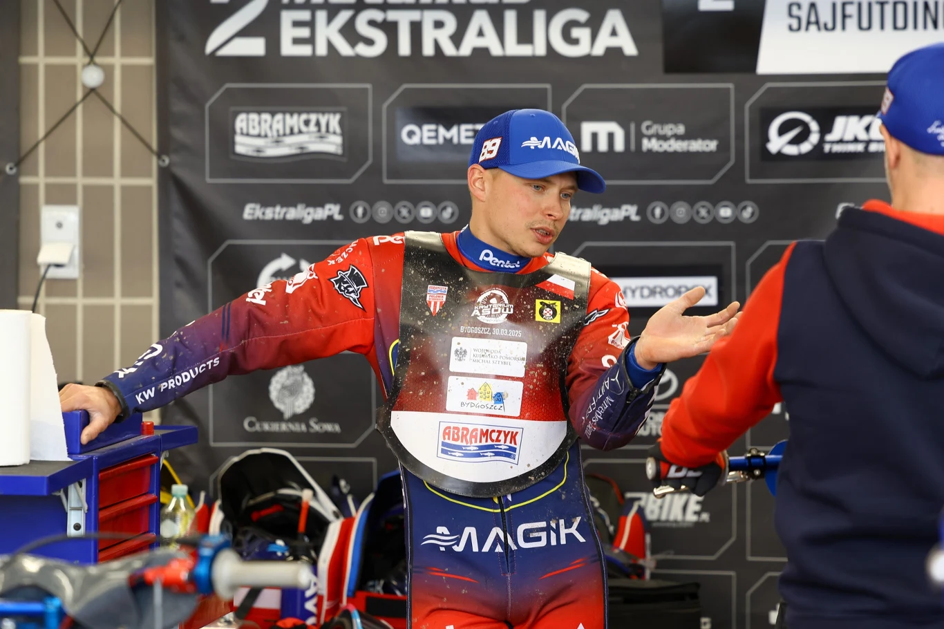 Zawodnik żużlowy w barwach drużyny stoi w parku maszyn podczas rozmowy z członkiem zespołu. W tle widoczne są logotypy sponsorów oraz motocykl żużlowy ustawiony obok nich.