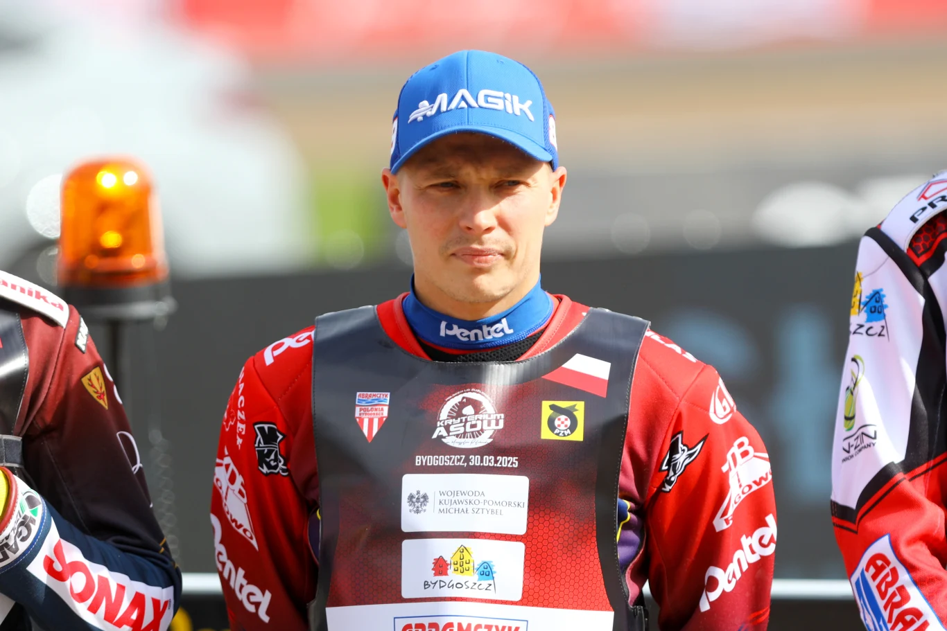 Zawodnik żużlowy w pełnym stroju z kaskiem na głowie, stojący obok innych uczestników na zawodach. Na jego kamizelce widoczne są logotypy sponsorów oraz godło narodowe.