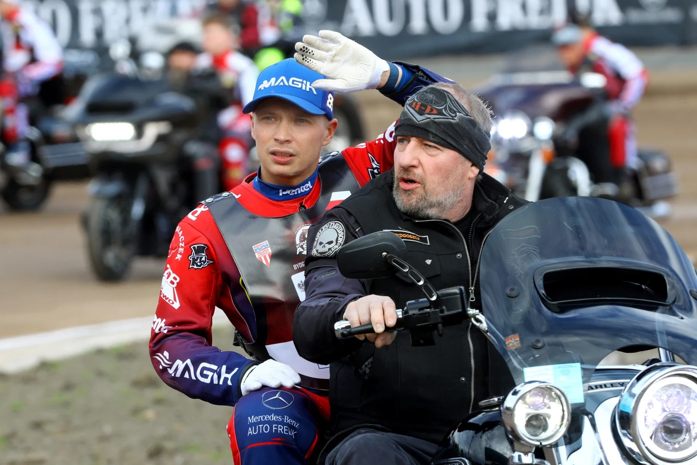Dwóch mężczyzn na motocyklu podczas wydarzenia sportowego. Jeden z nich ubrany w strój żużlowca, drugi w czarnej kurtce i opasce na głowie prowadzi maszynę. W tle inni uczestnicy oraz motocykle.