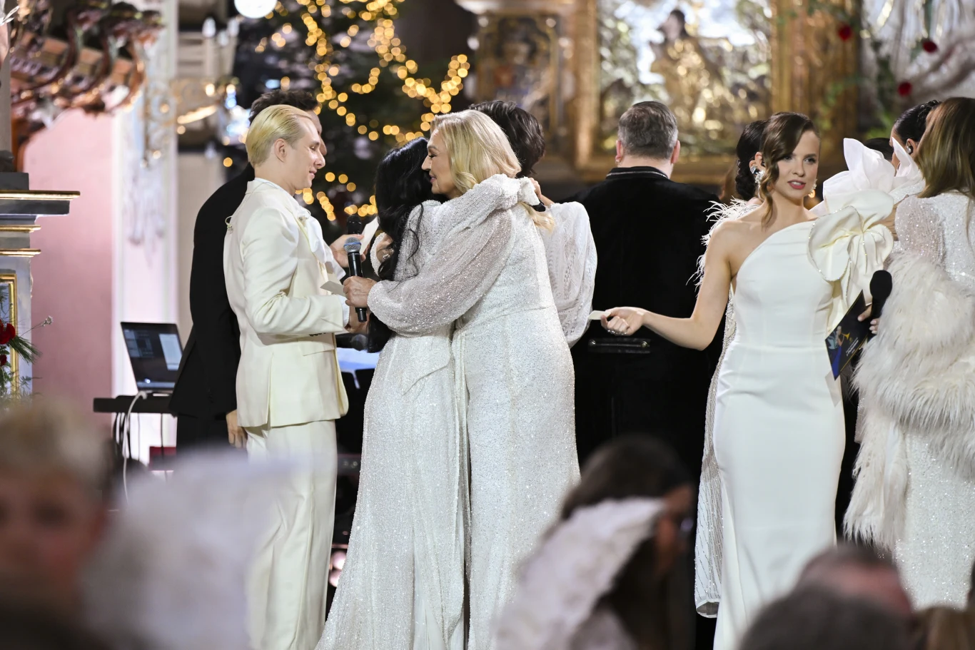 Grupa elegancko ubranych osób w białych strojach na scenie, część z nich trzyma mikrofony, niektórzy się przytulają, w tle ozdoby świąteczne i choinka.