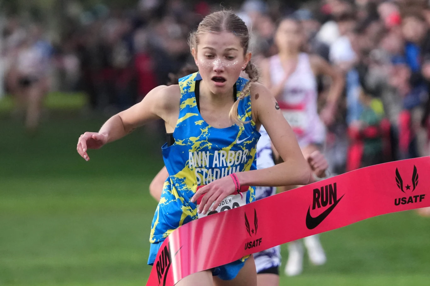 Natasza Dudek Młoda biegaczka w sportowym stroju z napisem Ann Arbor Stadium przekracza linię mety podczas zawodów biegowych, skupiona na finiszu, w tle rozmazana grupa innych zawodników.