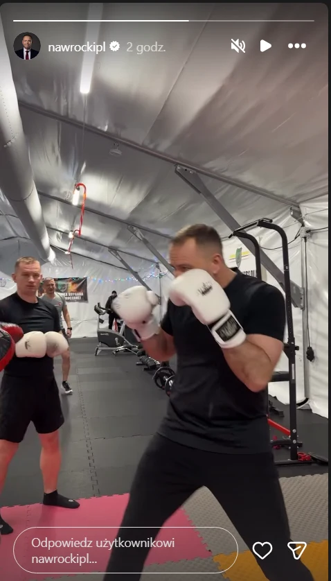 Dwóch mężczyzn w stroju sportowym trenuje boks w dużym, jasnym namiocie sportowym, wyposażonym w sprzęt do ćwiczeń. Główny uczestnik unosi rękawice bokserskie w pozycji obronnej, drugi mężczyzna stoi w tle w podobnej pozie.