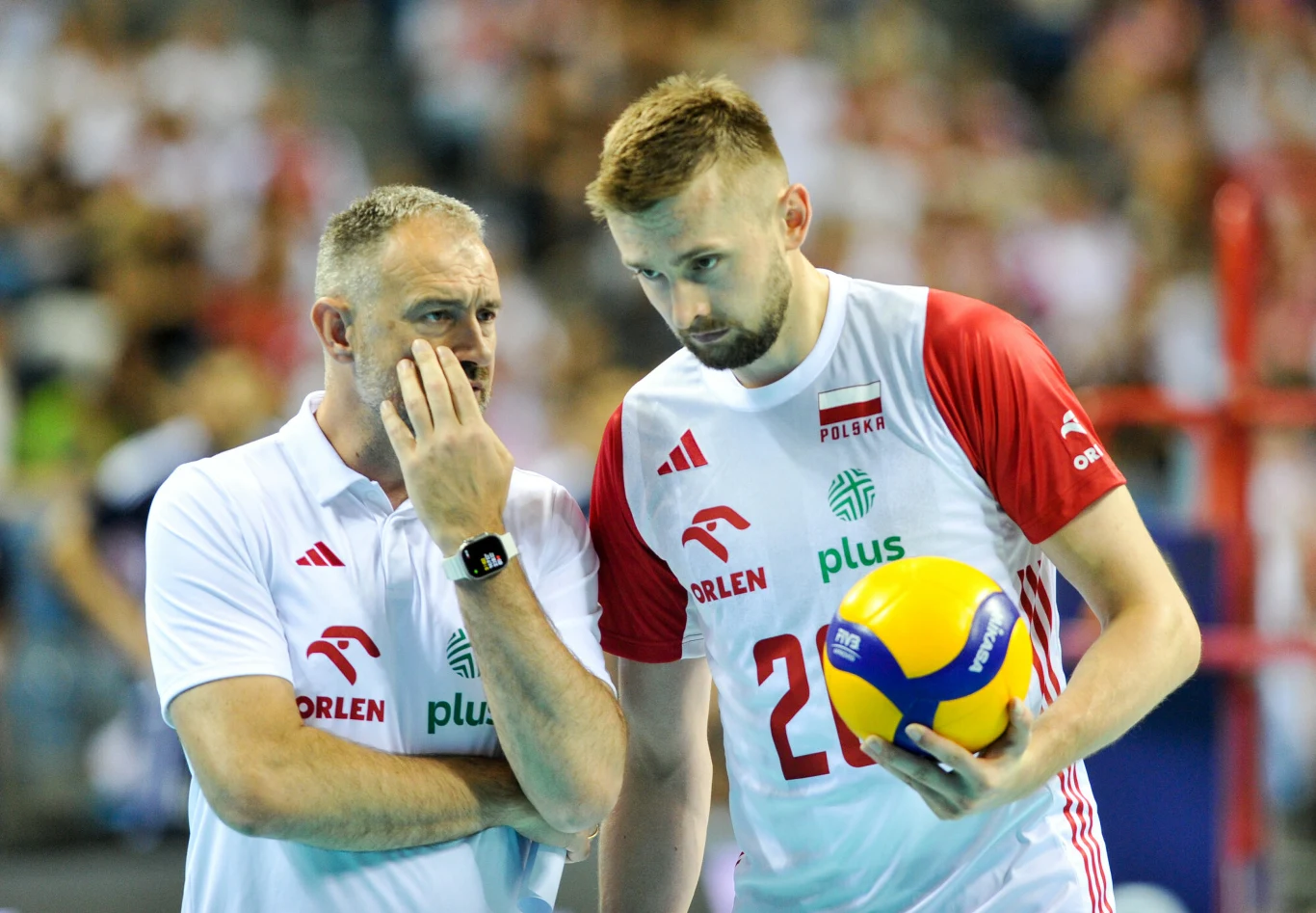 Trener i siatkarz drużyny narodowej Polski w trakcie rozmowy przy boisku, siatkarz trzyma piłkę do siatkówki, w tle rozmyta publiczność.