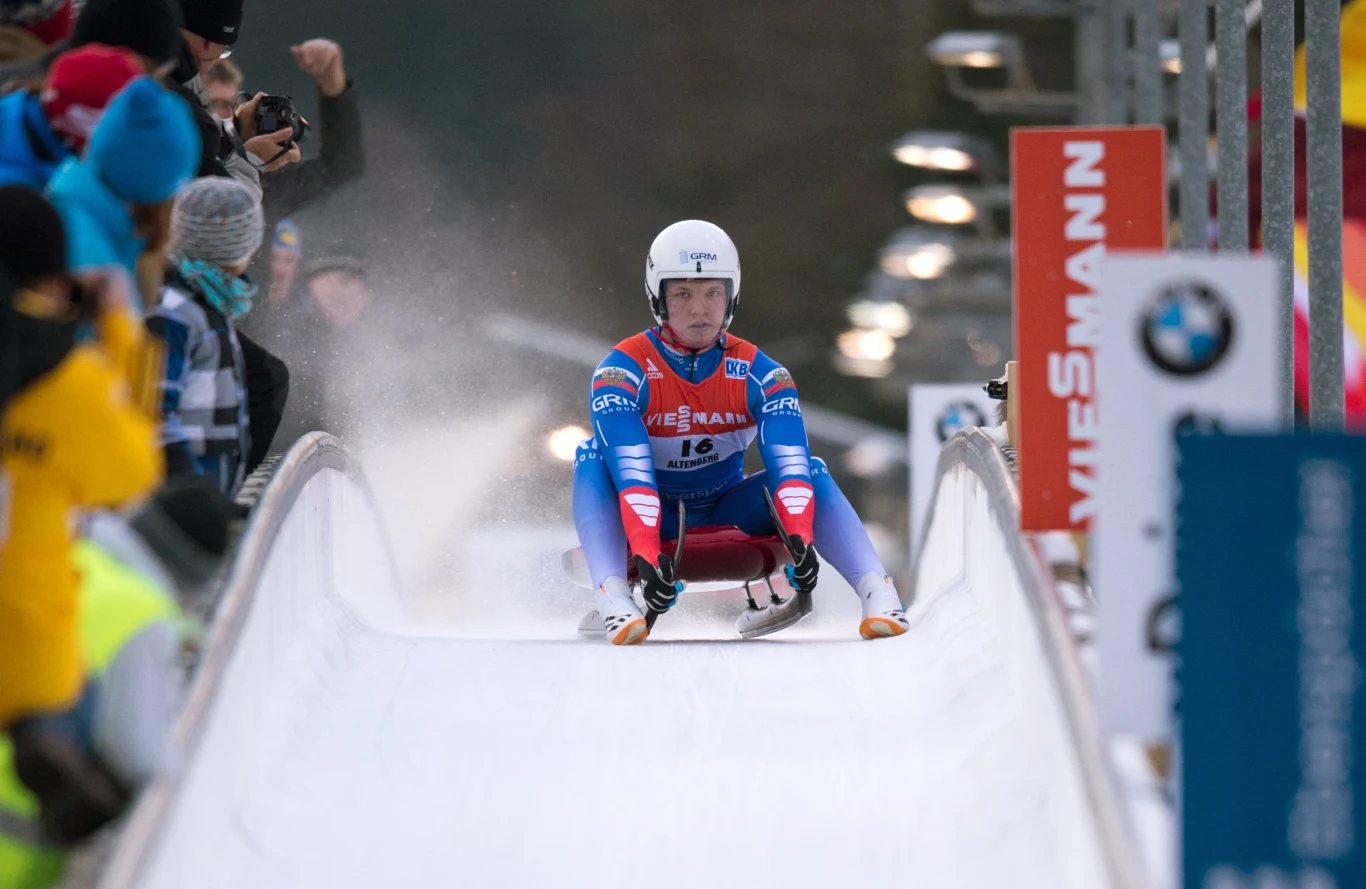 Zawodnik w kolorowym kombinezonie wykonuje zjazd na torze saneczkarskim, wokół zgromadzona jest grupa kibiców obserwujących rywalizację, w tle widoczne logo organizatorów zawodów.