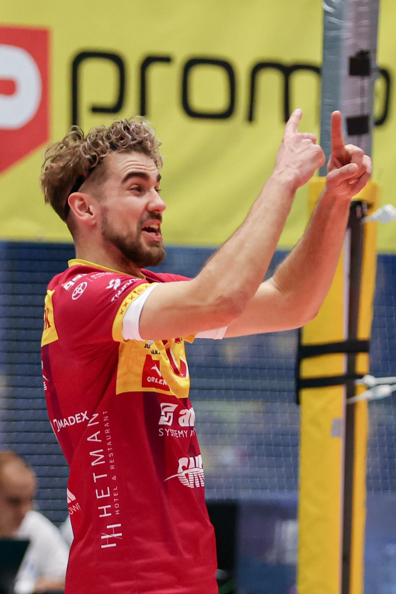 Siatkarz w czerwonym stroju z żółtymi akcentami gestykuluje rękami na tle siatkarskiego boiska i reklamowych banerów. Jego postawa i wyraz twarzy sugerują żywą komunikację podczas meczu sportowego.