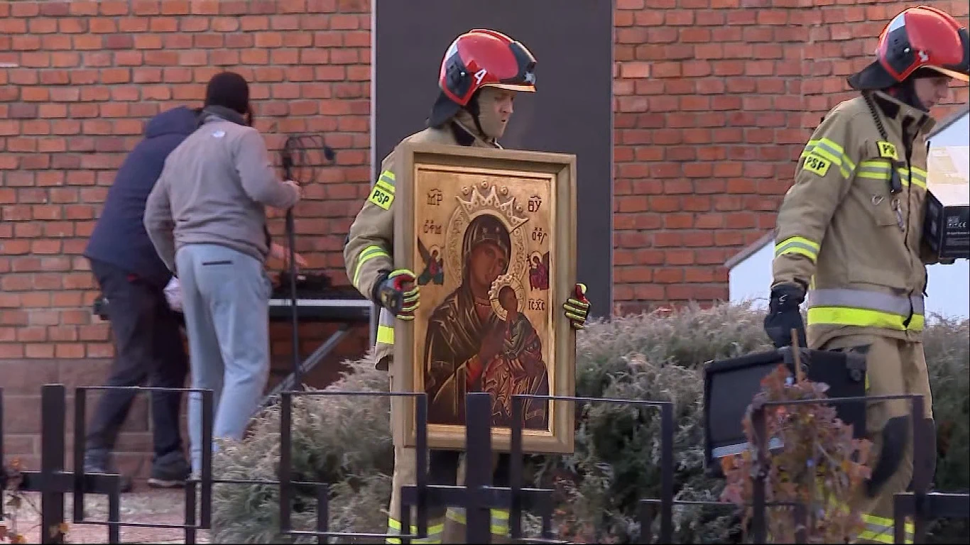 Strażak w pełnym umundurowaniu niesie dużą ikonę Matki Boskiej z Dzieciątkiem, w tle druga osoba w stroju strażaka oraz mężczyzna pracujący przy sprzęcie pod ceglaną ścianą.
