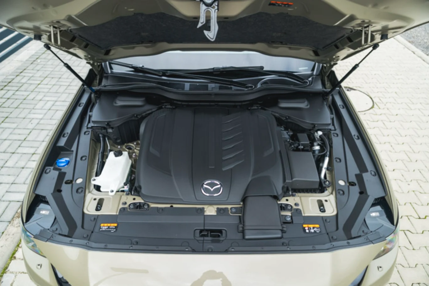 Silnik samochodu marki Mazda pod maską, z widocznym logo na pokrywie silnika. Elementy silnika otoczone plastikowymi osłonami, po bokach rozmieszczone kolorowe oznaczenia eksploatacyjne oraz zbiorniki płynów.