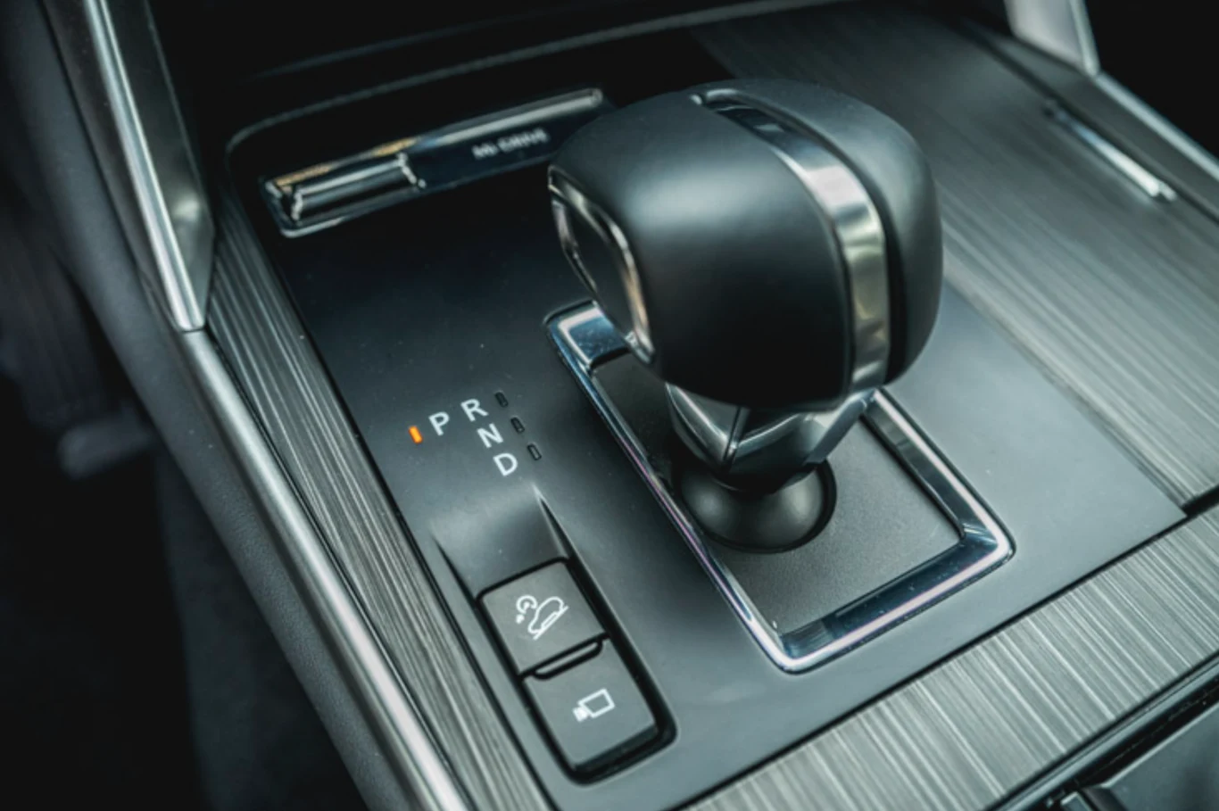 Nowoczesna dźwignia automatycznej skrzyni biegów w samochodzie, podświetlone oznaczenia trybów jazdy oraz przyciski funkcyjne na konsoli środkowej otoczone eleganckim wykończeniem.