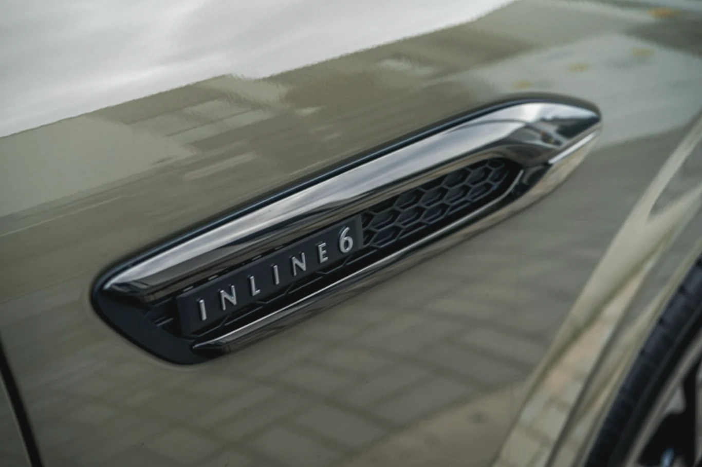 Detal auta z widocznym oznaczeniem 'INLINE 6' na błotniku, które wskazuje na typ silnika rzędowego o sześciu cylindrach, lakier powierzchni odbijający delikatnie otoczenie, elementy kratki wentylacyjnej.