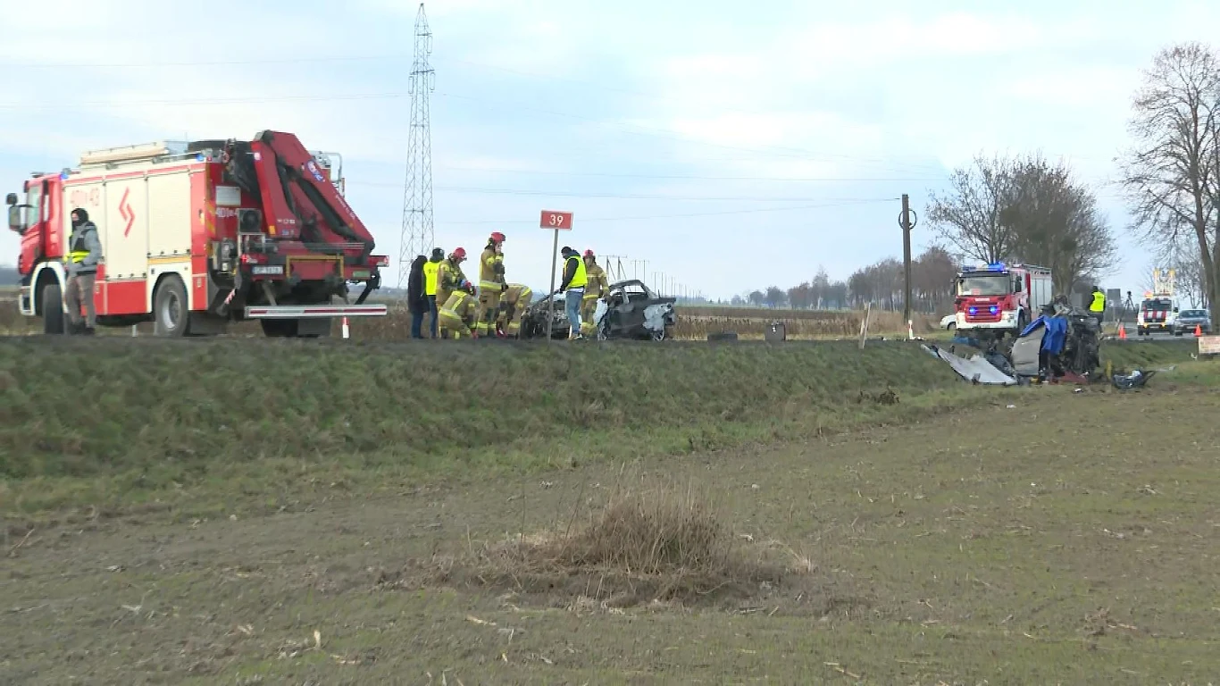 Służby ratunkowe pracują przy drodze obok rozbitego samochodu, używane są wozy strażackie, strażacy i ratownicy analizują miejsce wypadku, po jednej stronie rozciąga się pole, na drugim planie widać kolejne pojazdy służb.