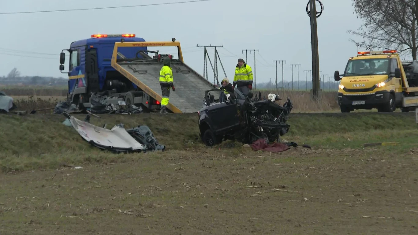 Rozbite auto po poważnym wypadku leżące przy poboczu, fragmenty wraku rozrzucone po okolicy, w tle dwie lawety i ratownicy ubrani w odblaskowe kamizelki przygotowujący się do usunięcia pojazdu, otoczenie stanowi wiejska droga i pola.