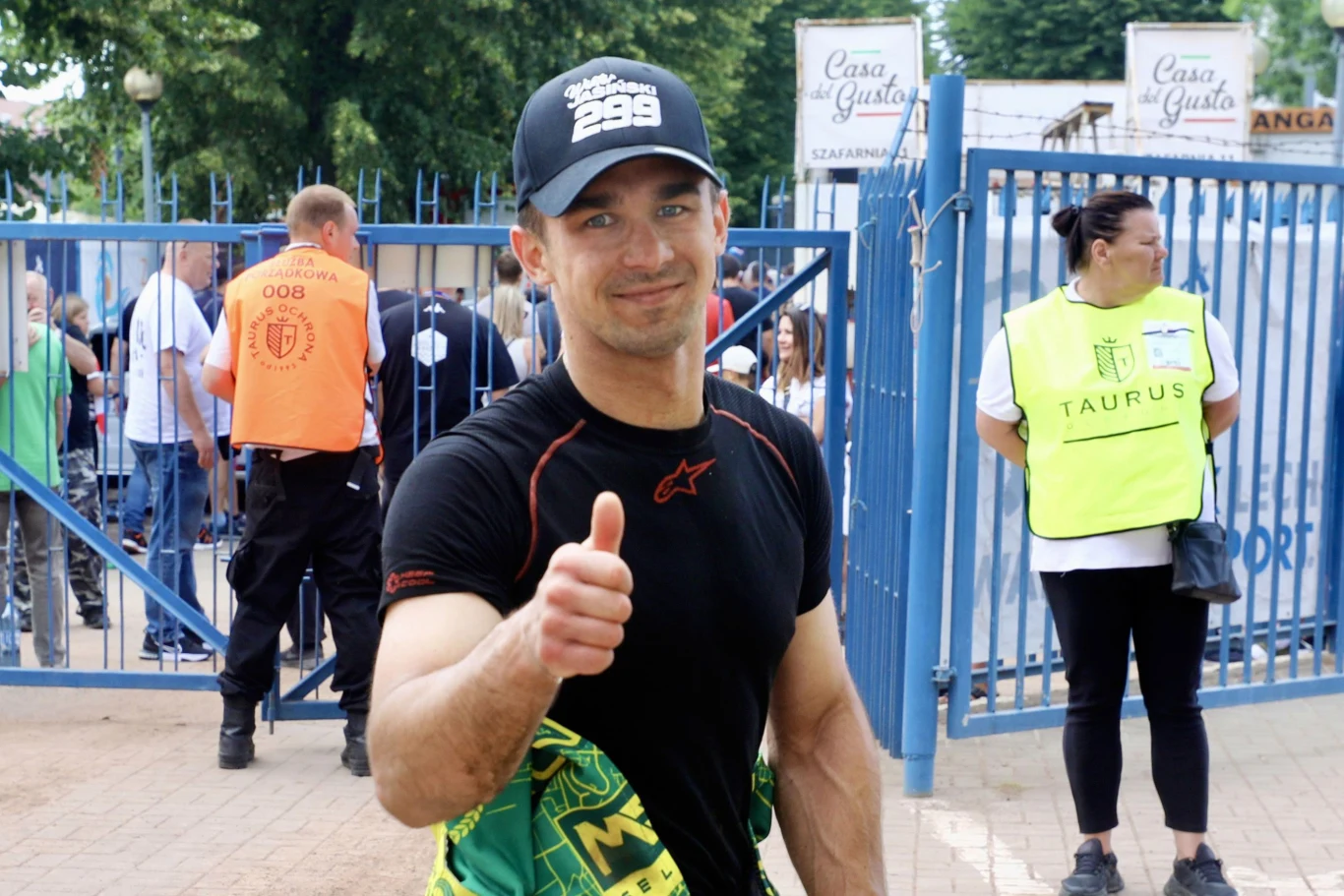 Wiktor Jasiński, zawodnik Pronergy Polonii Piła Mężczyzna w sportowym stroju i czapce z daszkiem uśmiecha się i pokazuje kciuk w górę, stojąc przed bramą, w tle widoczna ochrona oraz kilka osób uczestniczących w wydarzeniu plenerowym.
