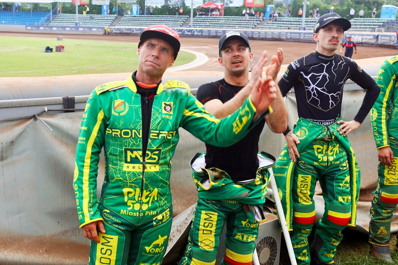 Od lewej: Norbert Kościuch, Wiktor Jasiński i Krzysztof Sadurski Grupa zawodników żużlowych w zielonych kombinezonach z oznaczeniami drużyny stoi na tle toru, jeden z nich wykonuje gesty rękami, wokół widoczne puste trybuny oraz barierki toru.