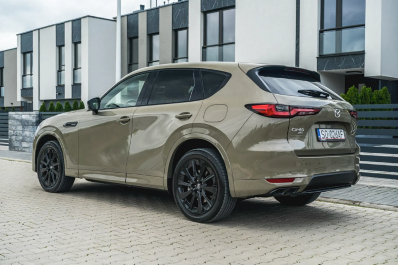 Brązowy SUV marki Mazda CX-60 zaparkowany na brukowanym podjeździe przed nowoczesnym budynkiem mieszkalnym, widoczne czarne felgi i przyciemniane szyby.