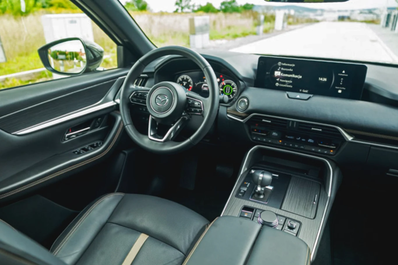Kokpit nowoczesnego samochodu z widocznym skórzanym wykończeniem, kierownicą z logotypem Mazda oraz dużym wyświetlaczem centralnym pokazującym system multimedialny. Deska rozdzielcza prezentuje czytelne wskaźniki oraz nowoczesną konsolę środkową.