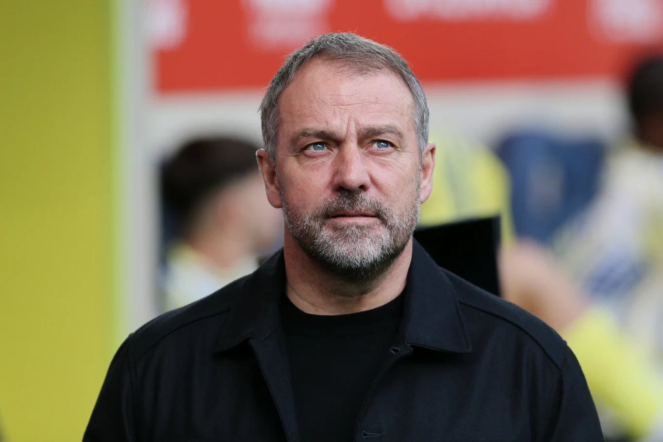 Mężczyzna o siwiejących włosach i brodzie, ubrany w ciemną kurtkę, stoi na tle rozmazanego żółto-czerwonego tła, patrząc poważnie przed siebie.