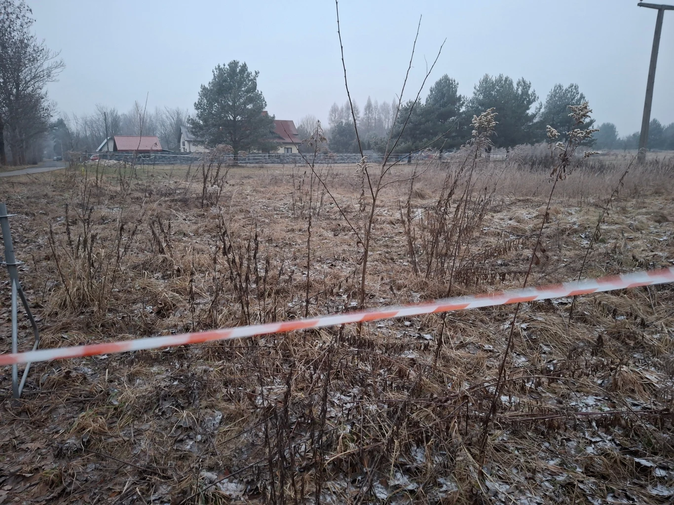 Pusty, zaśnieżony teren otoczony taśmą ostrzegawczą z widocznymi suchymi roślinami, w tle zabudowania i drzewa na tle pochmurnego nieba.