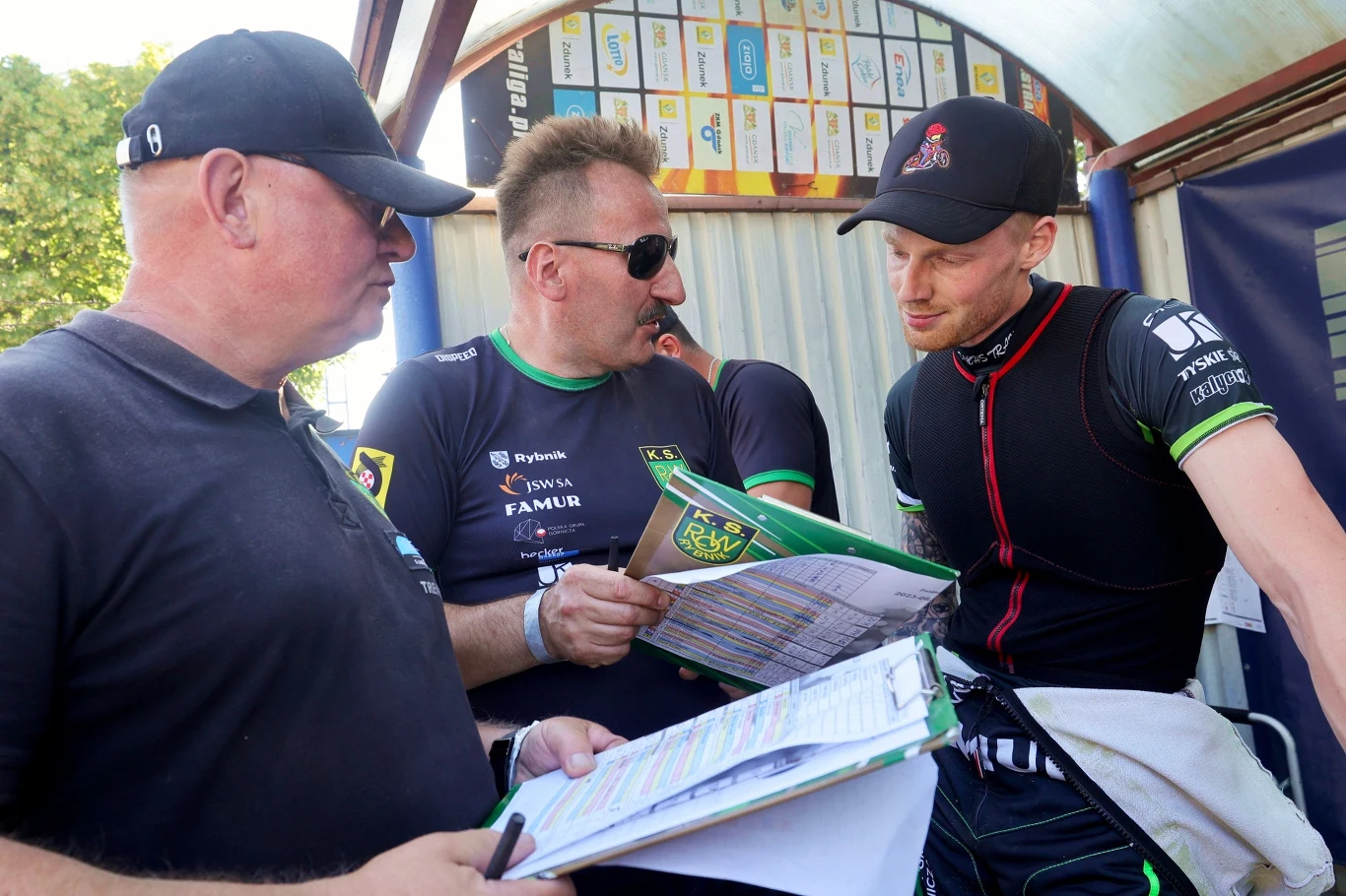 Krzysztof Mrozek i Patrick Hansen w 2023 roku Trzech mężczyzn omawia dokumenty i notatki podczas wydarzenia sportowego, w tle widoczne plansze z logotypami sponsorów. Jeden z mężczyzn nosi strój sportowy i czapkę z daszkiem, pozostali ubrani są w czarne koszulki z nadrukami.