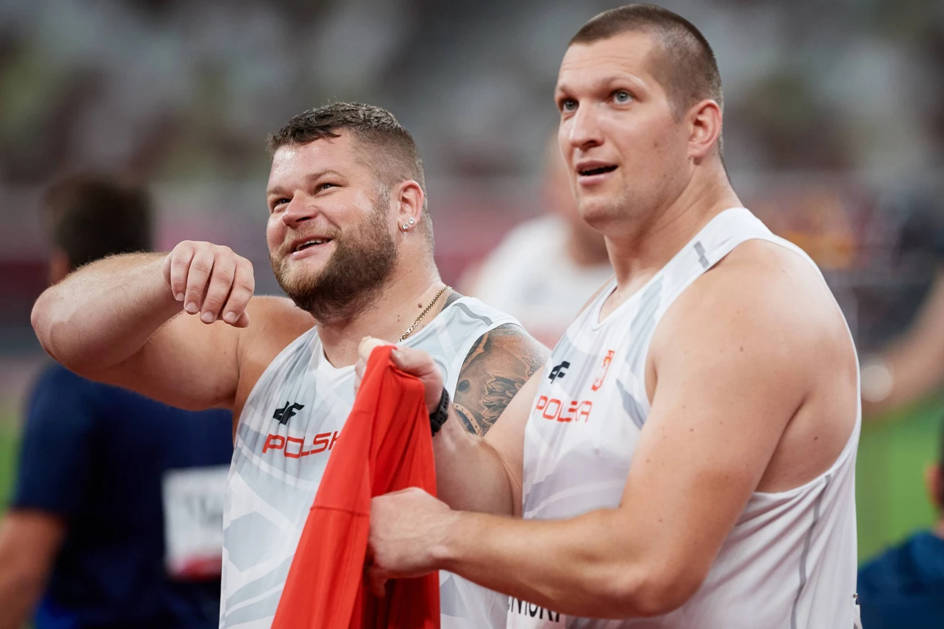 Dwóch sportowców w biało-czerwonych strojach z napisem Polska, w wyraźnie dobrym nastroju oraz z polską flagą w rękach, znajdują się na stadionie lekkoatletycznym.