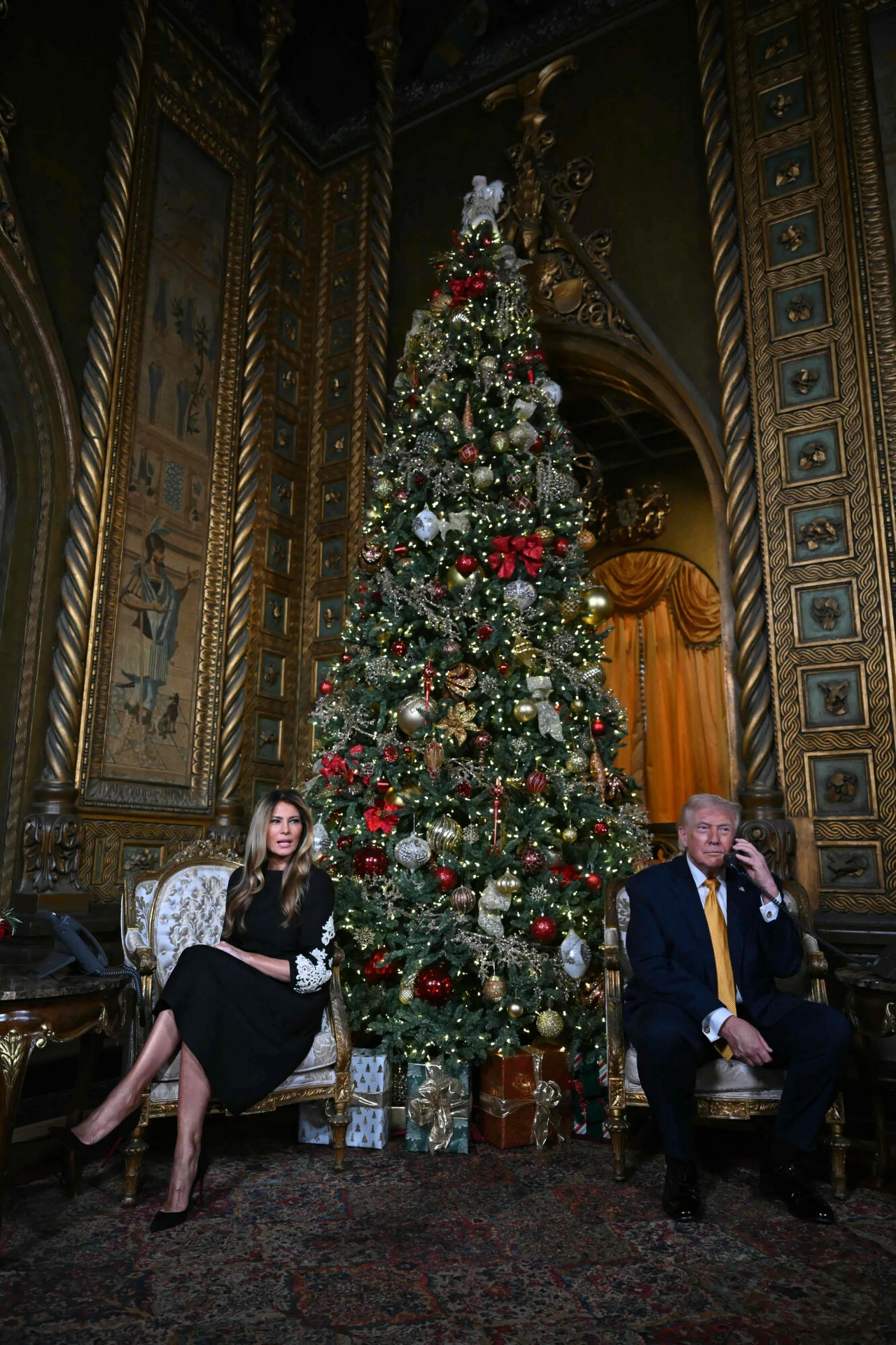 Bogato udekorowana choinka świąteczna ustawiona w luksusowym, złotym wnętrzu z dwoma osobami siedzącymi po obu stronach drzewa, zaawansowane zdobienia ścian i mebli, prezenty pod choinką, eleganckie ubrania obecnych osób.