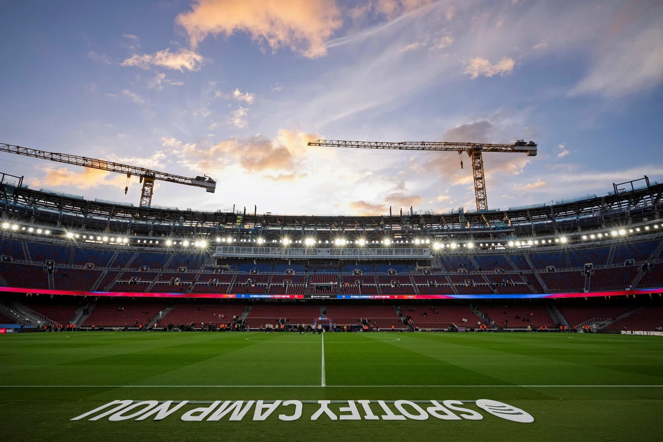 Rozległy stadion piłkarski z widocznymi trybunami, otoczony dwoma dużymi dźwigami budowlanymi na tle wieczornego nieba, centralnie umieszczone boisko, napisy na murawie.