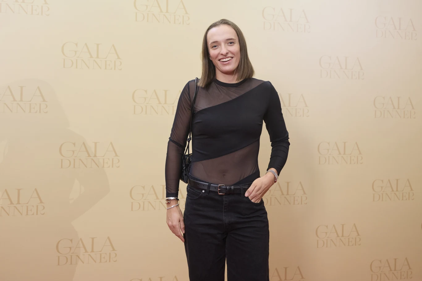 Kobieta ubrana w czarną, częściowo prześwitującą bluzkę oraz czarne spodnie stoi na tle ściany z powtarzającym się napisem GALA DINNER, uśmiechnięta, z rozpuszczonymi włosami, pozująca w stylu eventowym.