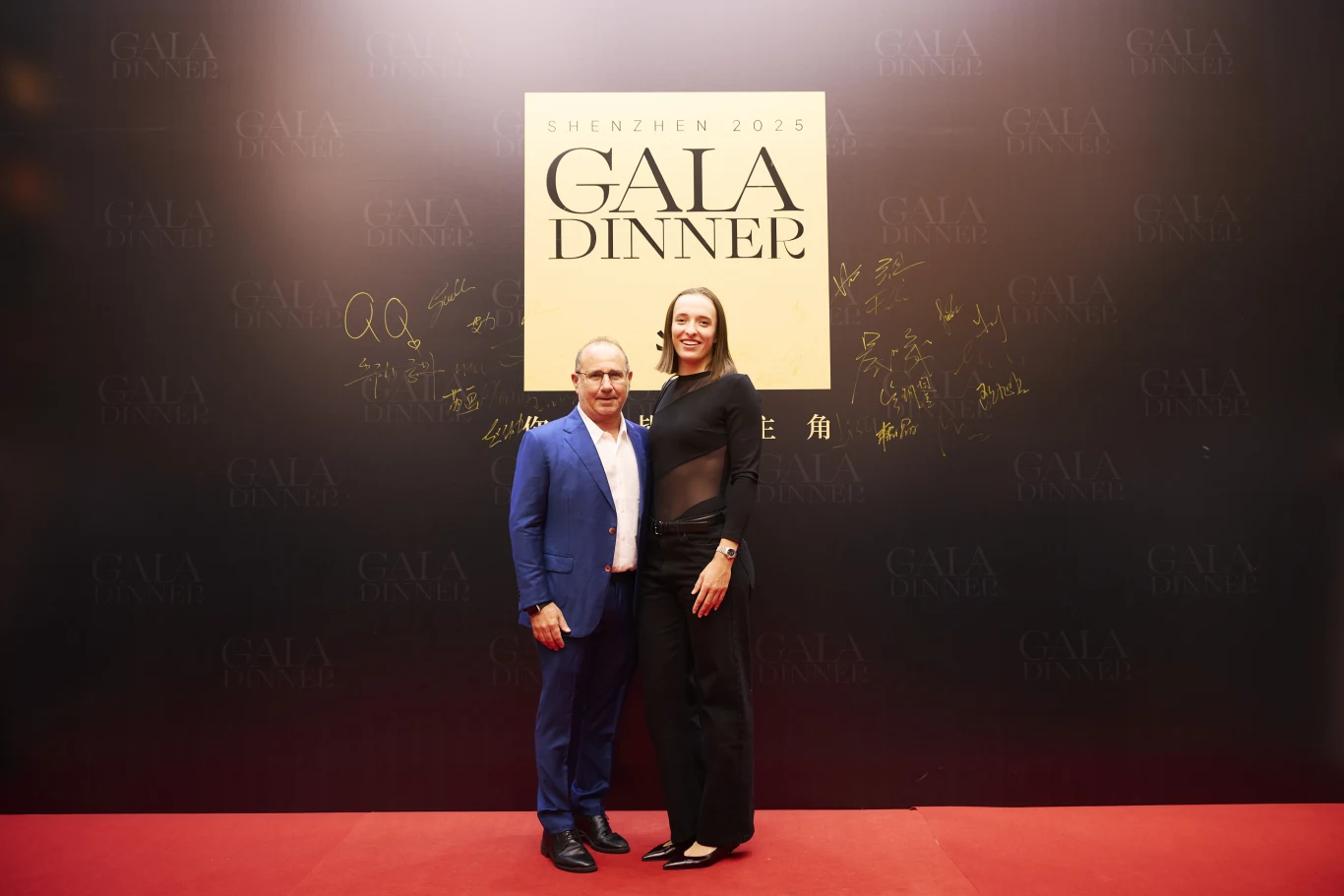 Dwoje elegancko ubranych ludzi pozuje na tle czarnej ścianki z napisem Gala Dinner Shenzhen 2025; otacza ich czerwony dywan i złote podpisy na tle ściany.