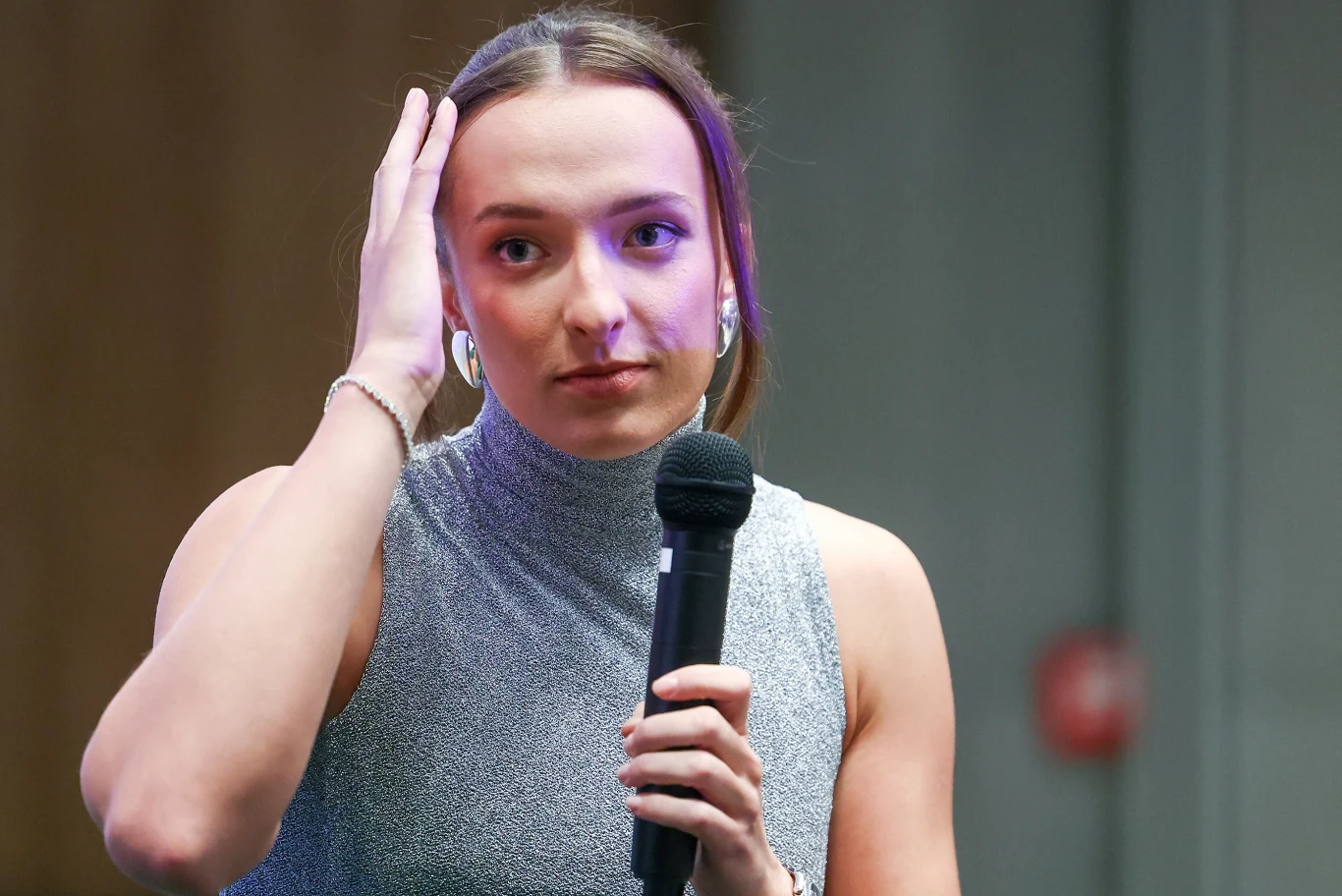 Młoda kobieta w jasnoszarej bluzce trzyma mikrofon w jednej ręce, drugą dotyka włosów. Skupiony wyraz twarzy, neutralne tło.