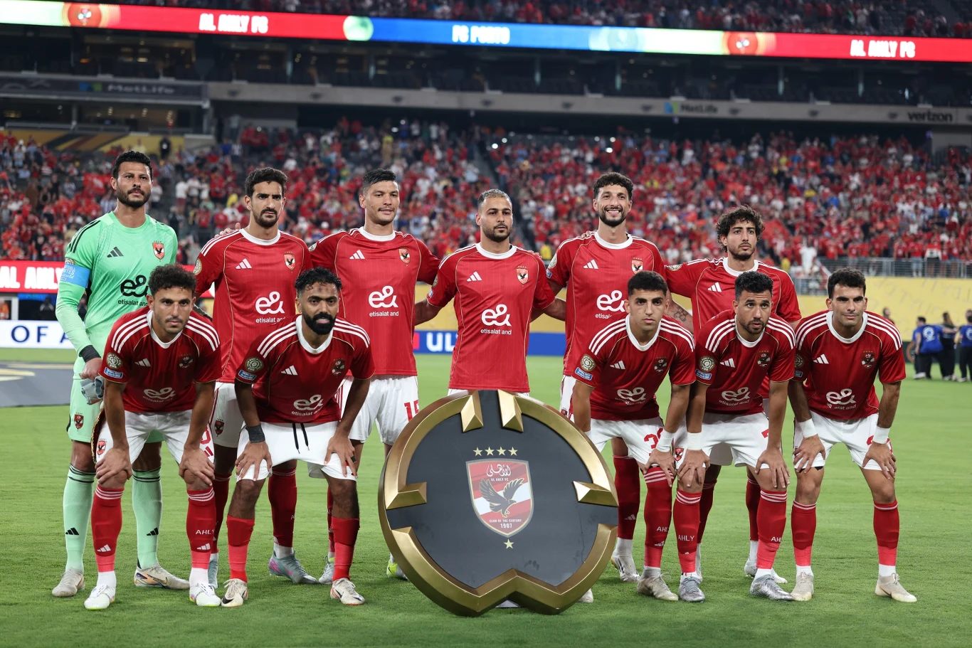 Piłkarze drużyny ubrane w czerwone stroje stojące na murawie stadionu, trzymający duży medal lub tarczę symbolizującą trofeum. W tle liczna publiczność na trybunach.